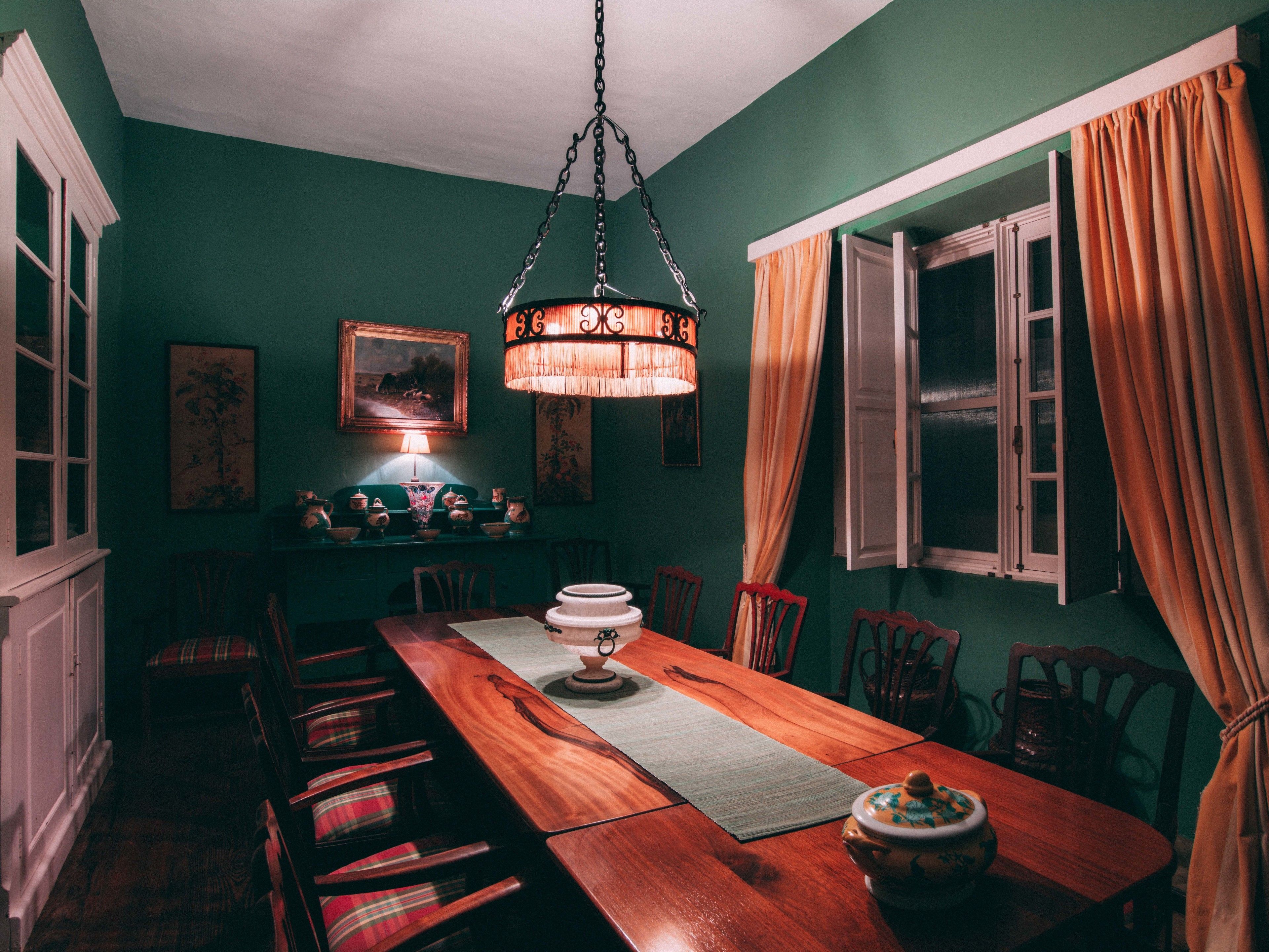 A dining room with a wooden table, chairs, a hanging light fixture, and a window with curtains.