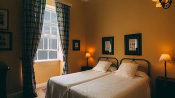 A room with two beds, a window with plaid curtains, and wall-mounted lamps.