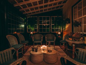 Um quarto acolhedor com móveis de vime, uma mesa redonda central decorada com velas e grandes janelas com plantas.