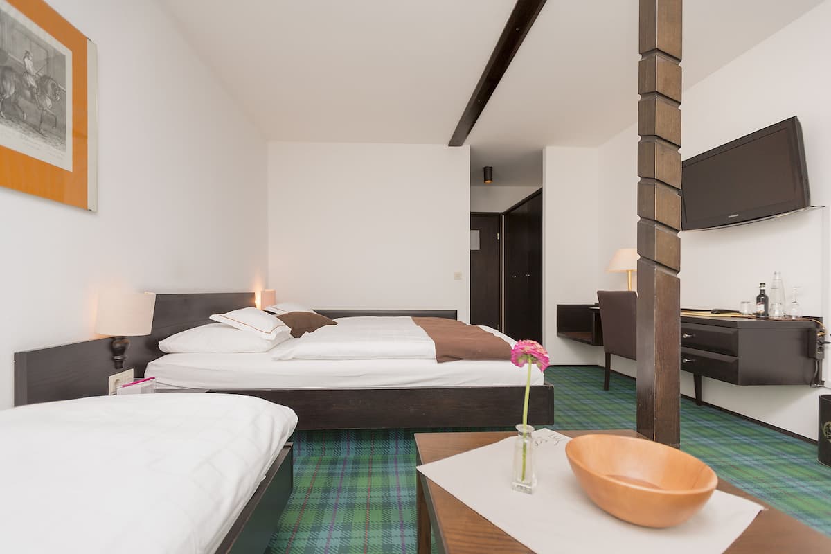 Quarto de hotel com duas camas, uma mesa de trabalho, televisão e banheiro ao fundo.
