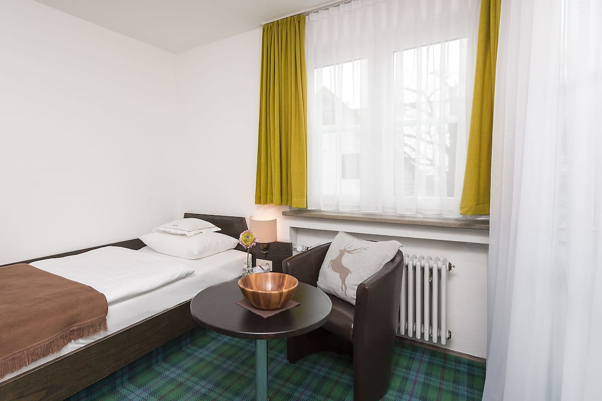 Quarto de hotel com duas camas de solteiro, uma mesinha com abajur, radiador e janela com cortinas verdes.