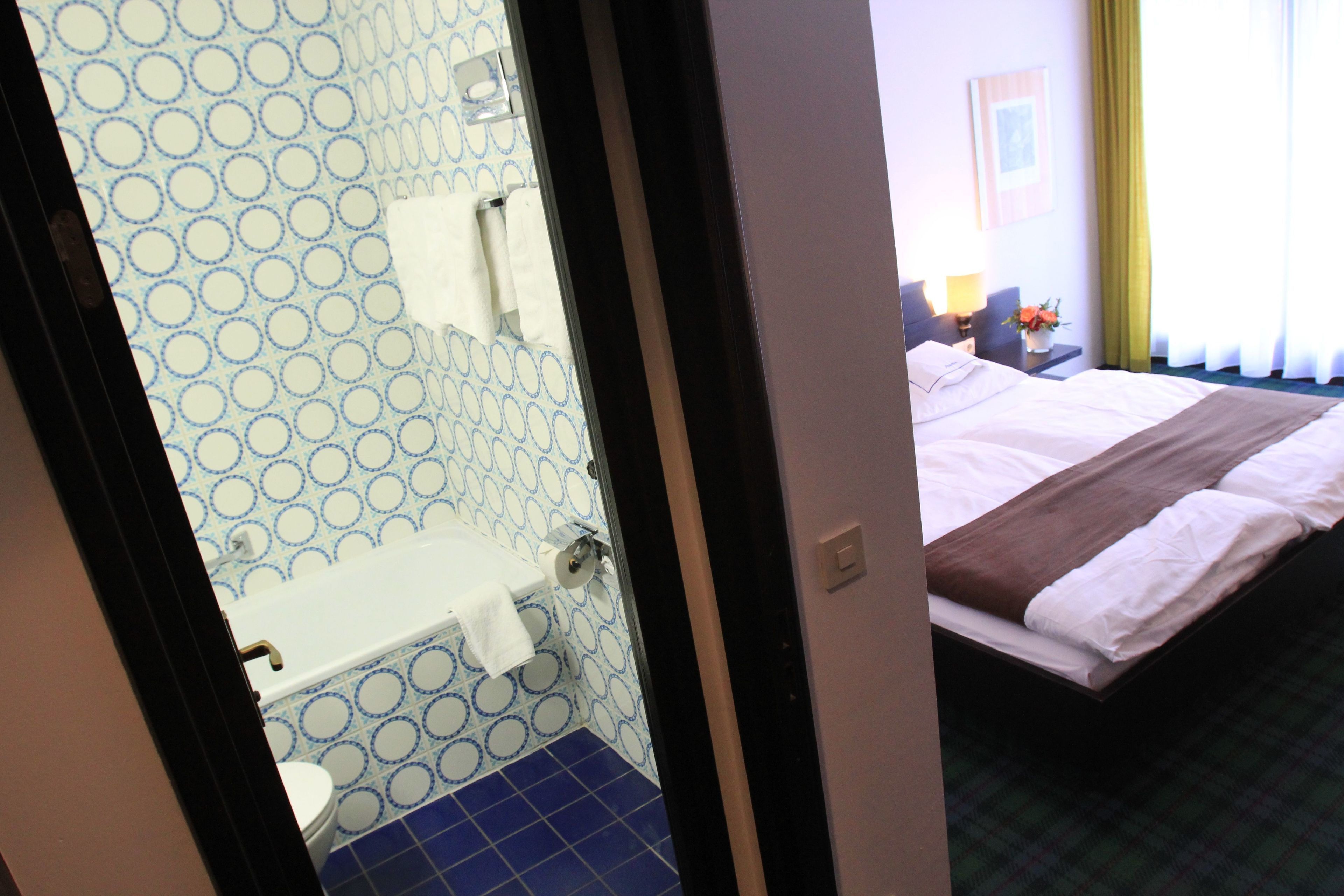A hotel room with a bathroom featuring blue and white patterned tiles, a white toilet, and a sink with a towel. The bed has a brown headboard and white bedding.