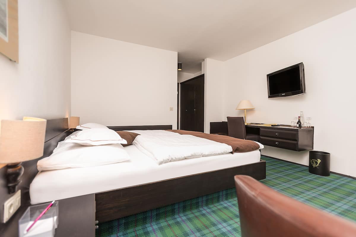 Quarto de hotel com cama, mesa de cabeceira, abajur, televisão e pequena escrivaninha com cadeira.