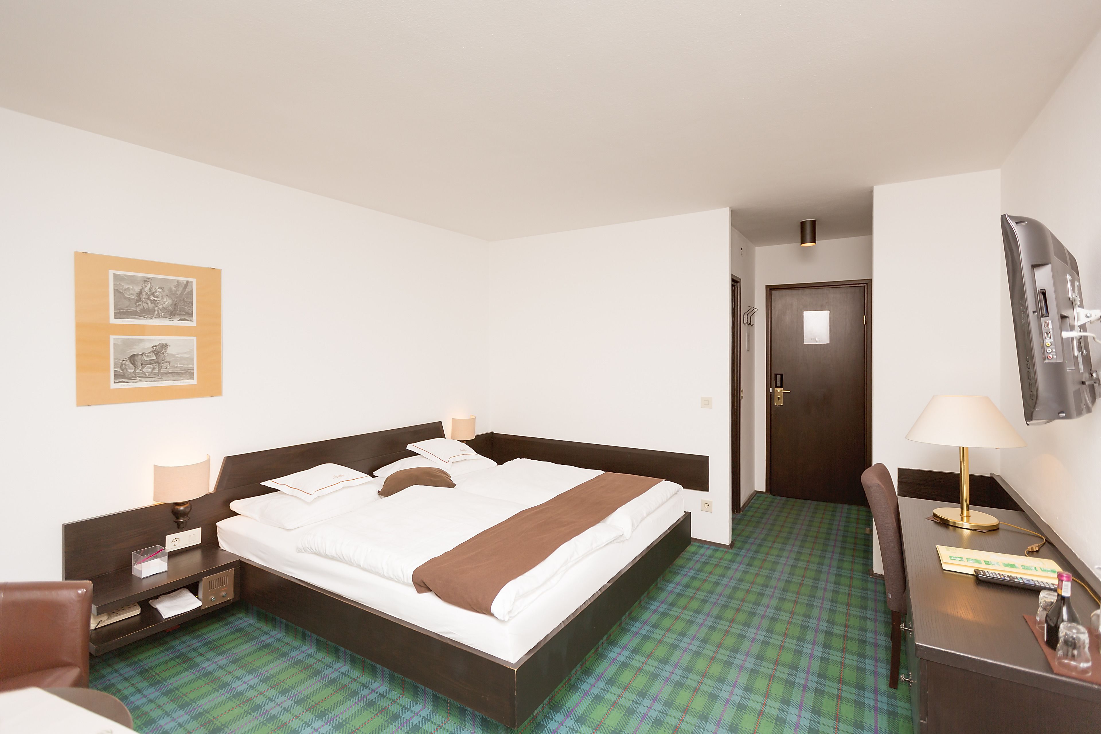 Habitación de hotel con cama, mesitas de noche, escritorio, silla, lámpara y puerta.