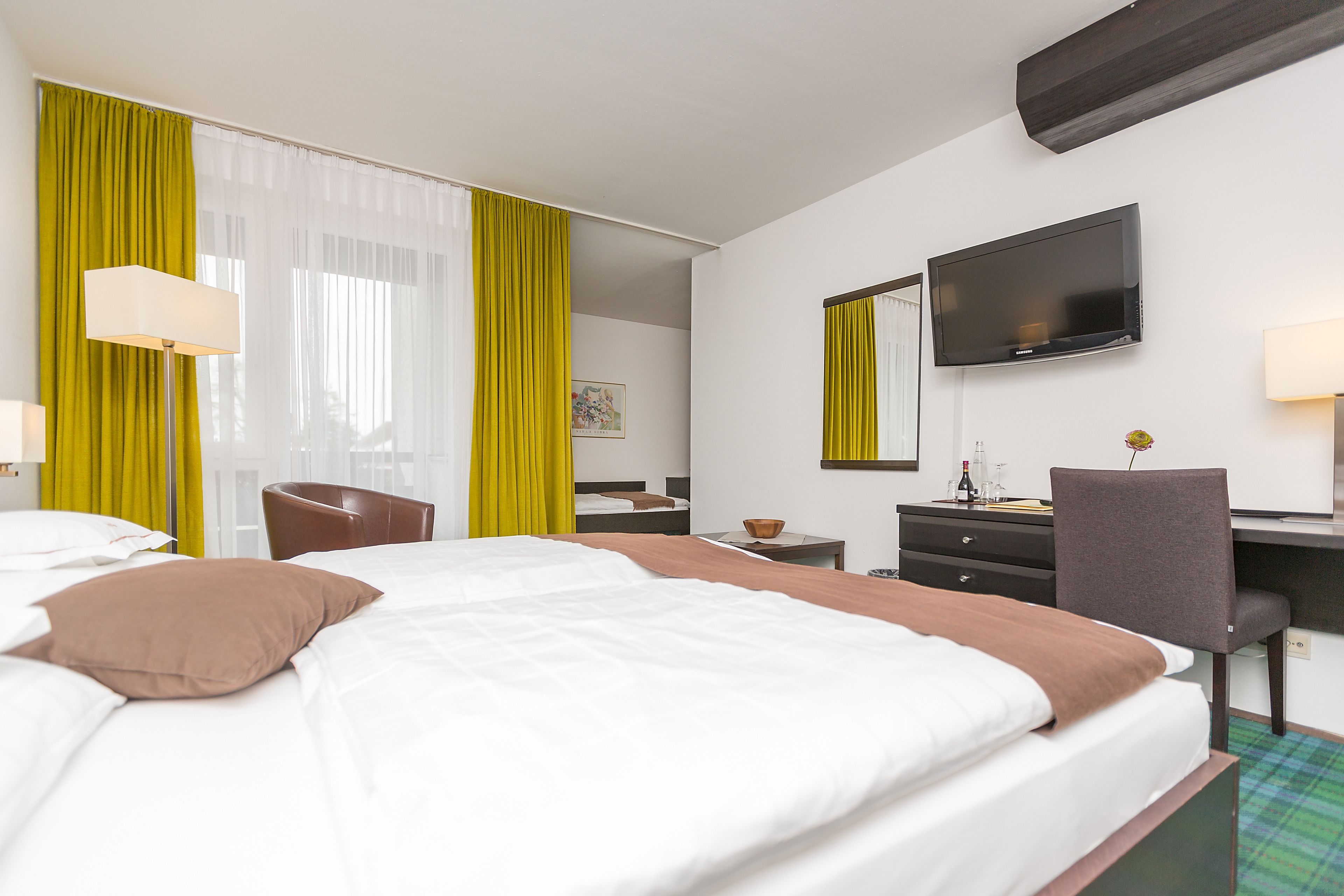 Habitación de hotel con cama, escritorio, silla, televisor y ventana con cortinas.