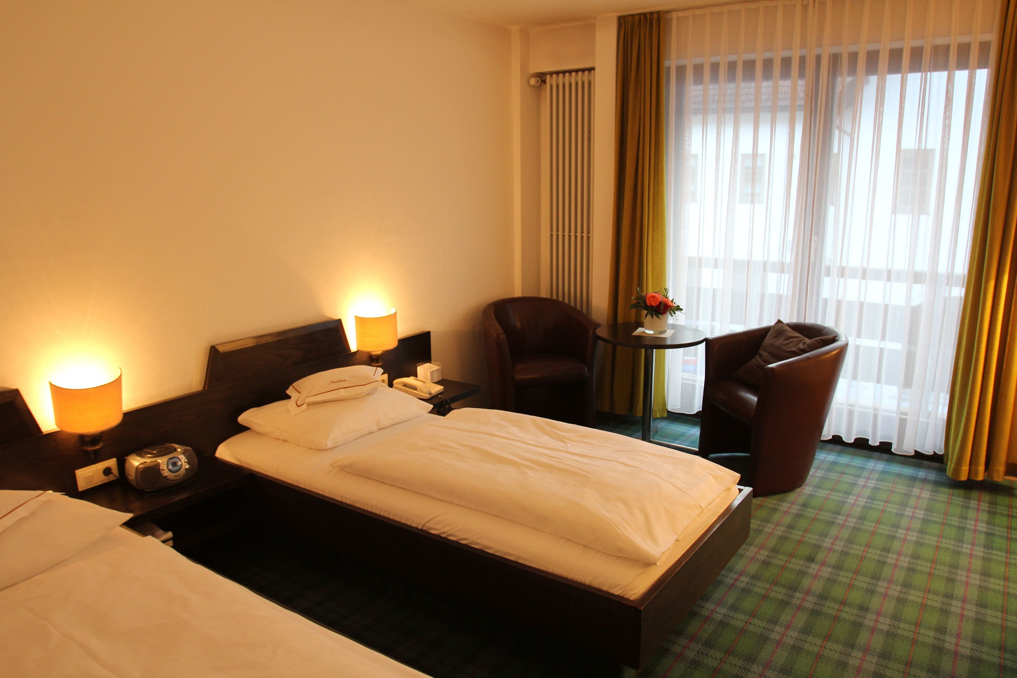 A hotel room with two beds, a desk, a chair, and a view of the outdoors through large windows.