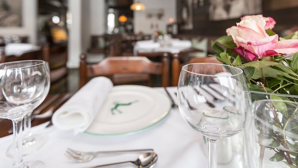 Une table dressée pour deux dans un restaurant, avec une nappe blanche, une assiette verte et blanche, et un vase contenant une fleur rose.