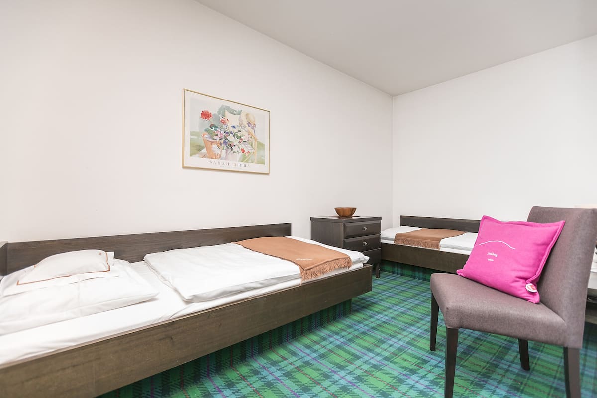 Quarto de hotel com duas camas de solteiro, uma cadeira, uma mesinha com uma tigela e um quadro na parede.