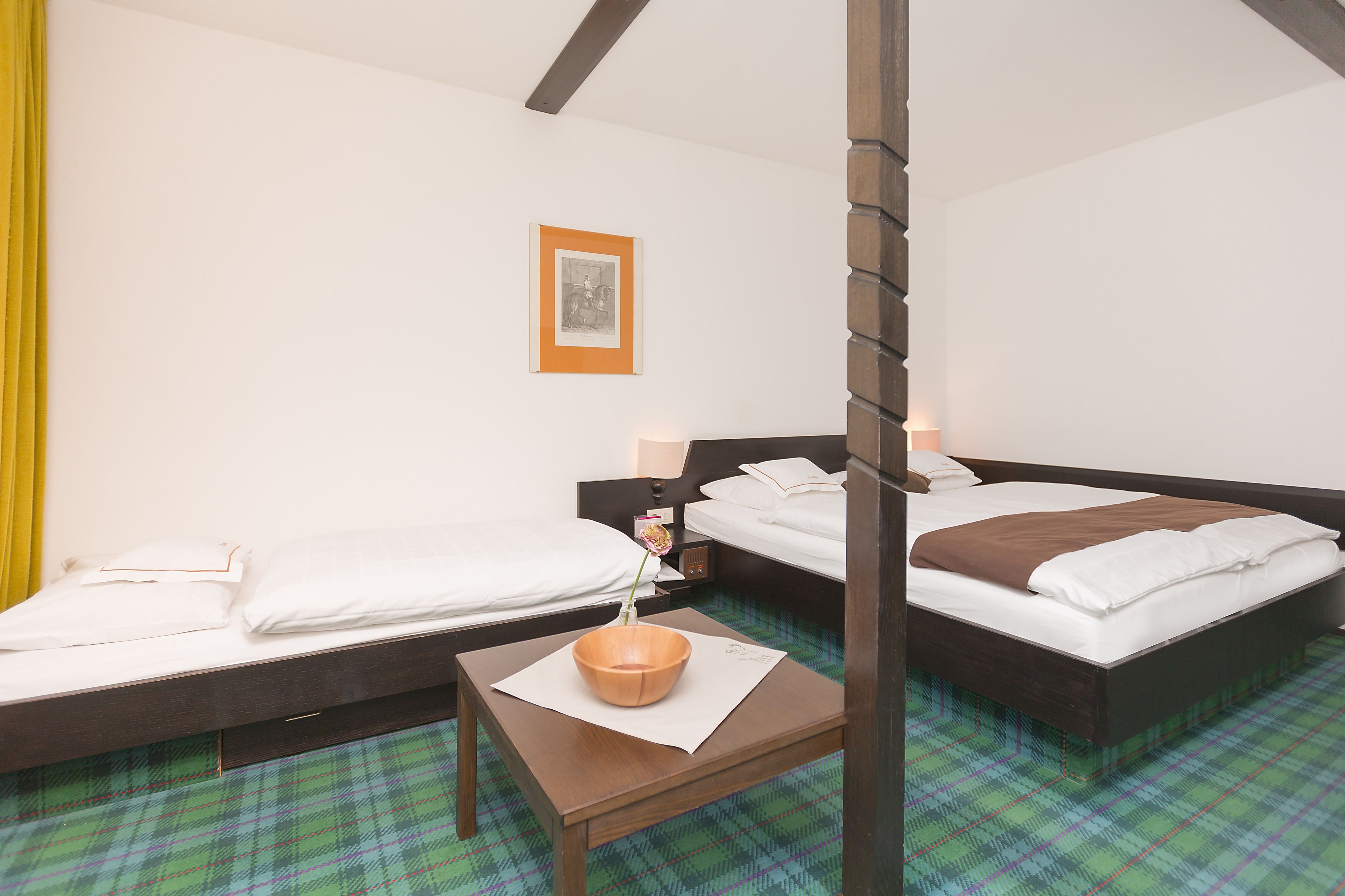 A hotel room with two single beds, a wooden nightstand with a vase, and a green plaid carpet.