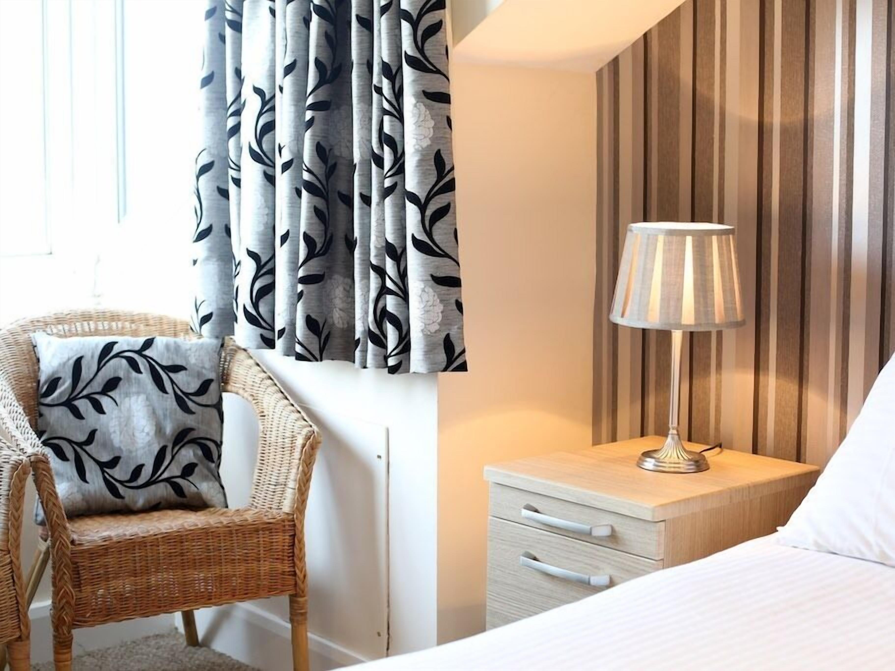 Een hotelkamer met een rieten stoel, een nachtkastje met een lamp, en een bed met wit beddengoed.
