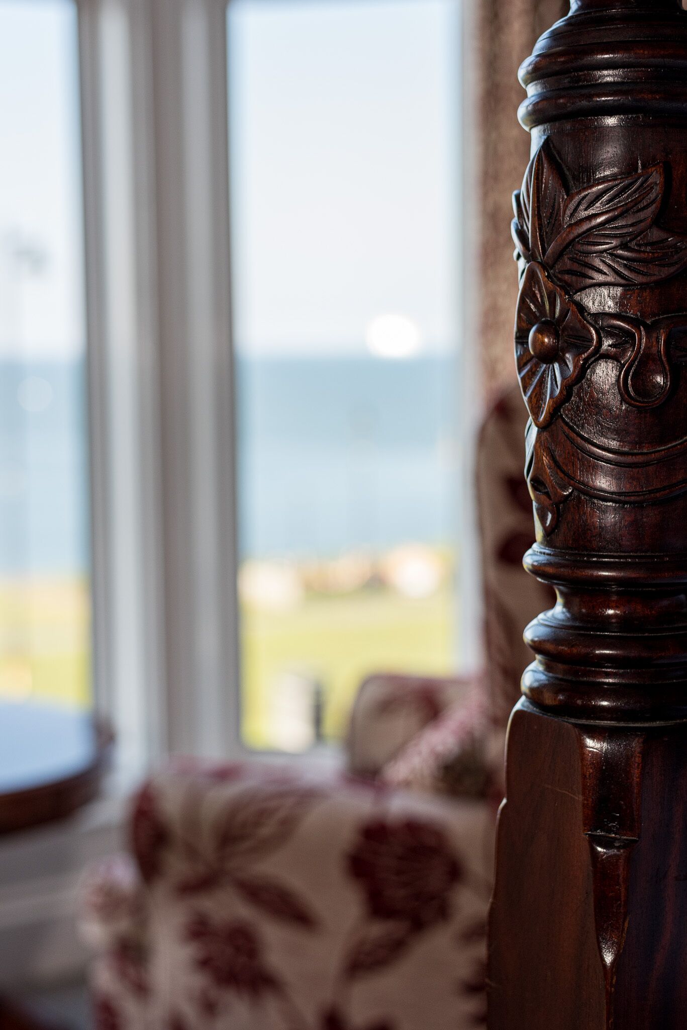 A close-up of a wooden chair leg with intricate carvings, set against a blurred background of a window with a view of a landscape.