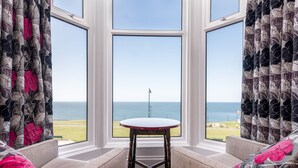 A room with a view of the sea through large windows, a round table, and floral curtains. - The Riviera Guest House (Whitby)