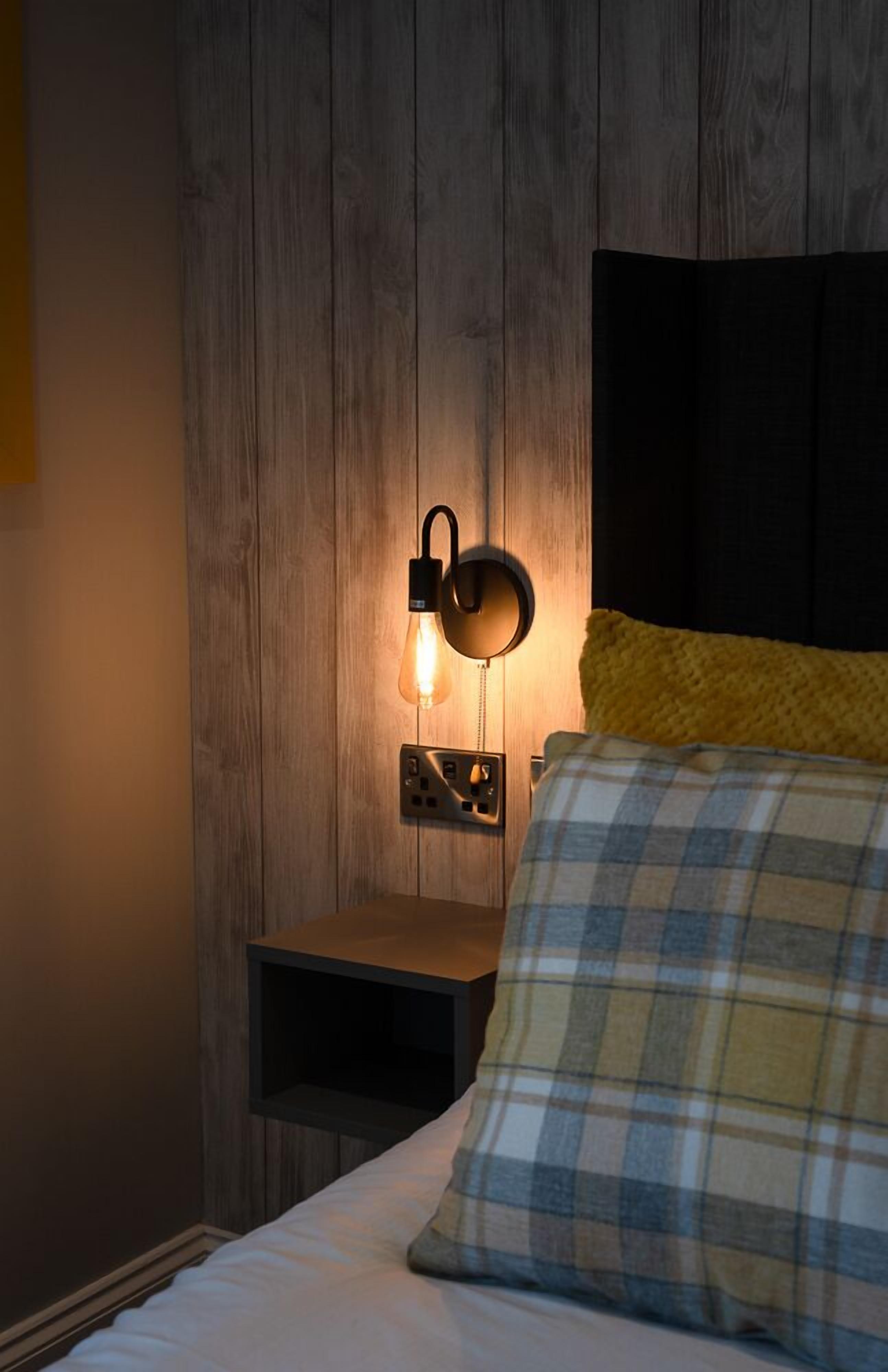 Une chambre avec un mur en bois, une table de chevet, une lampe et un lit recouvert d’une couverture à carreaux.