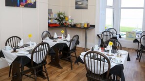 A dining area with wooden chairs and tables, a buffet counter with food and drinks, and a view of the sea through large windows. - The Riviera Guest House (Whitby)