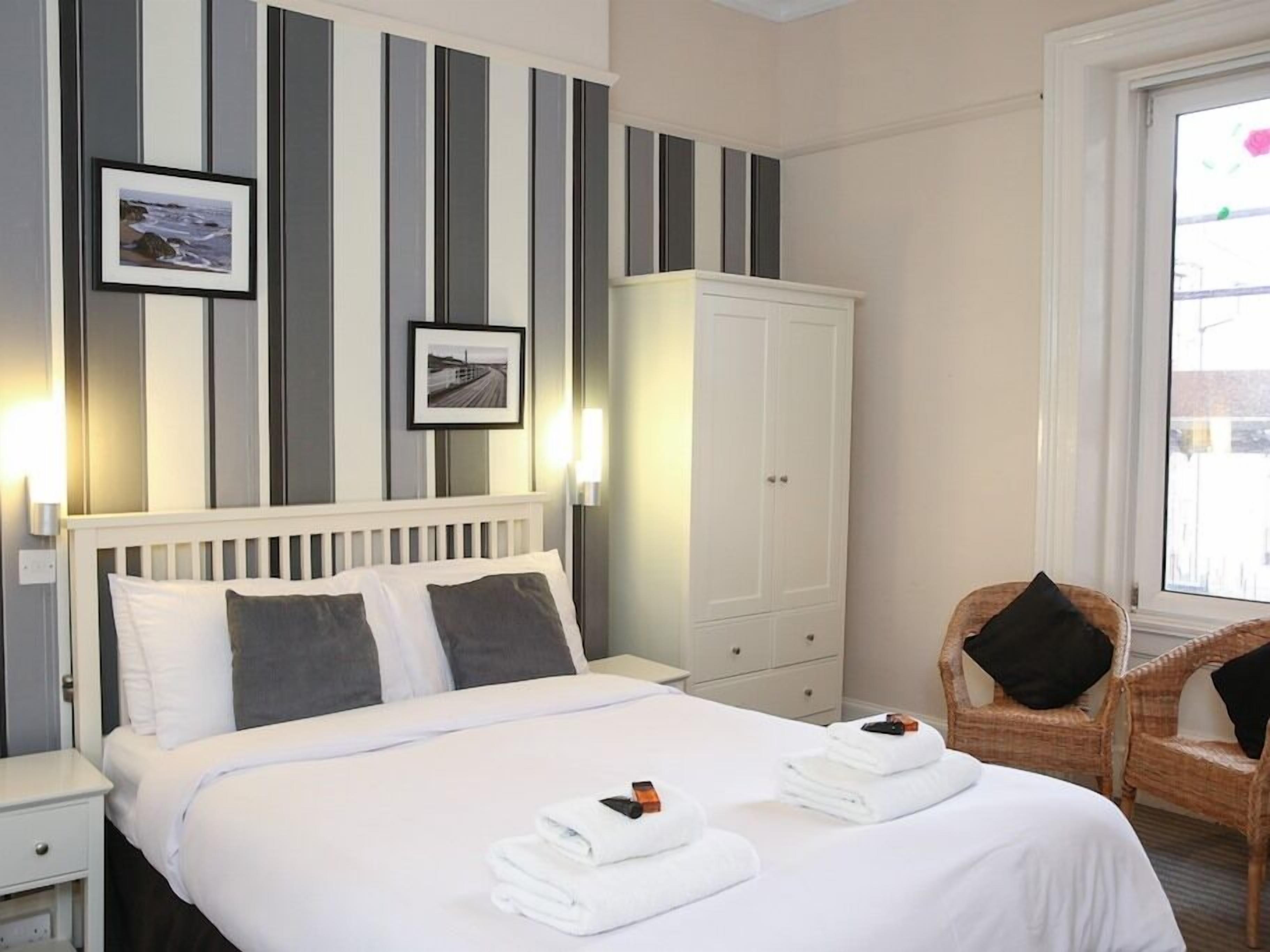 A hotel room with a bed, white headboard, striped wall decor, two framed pictures, a white wardrobe, a wicker chair, and a window with a view.