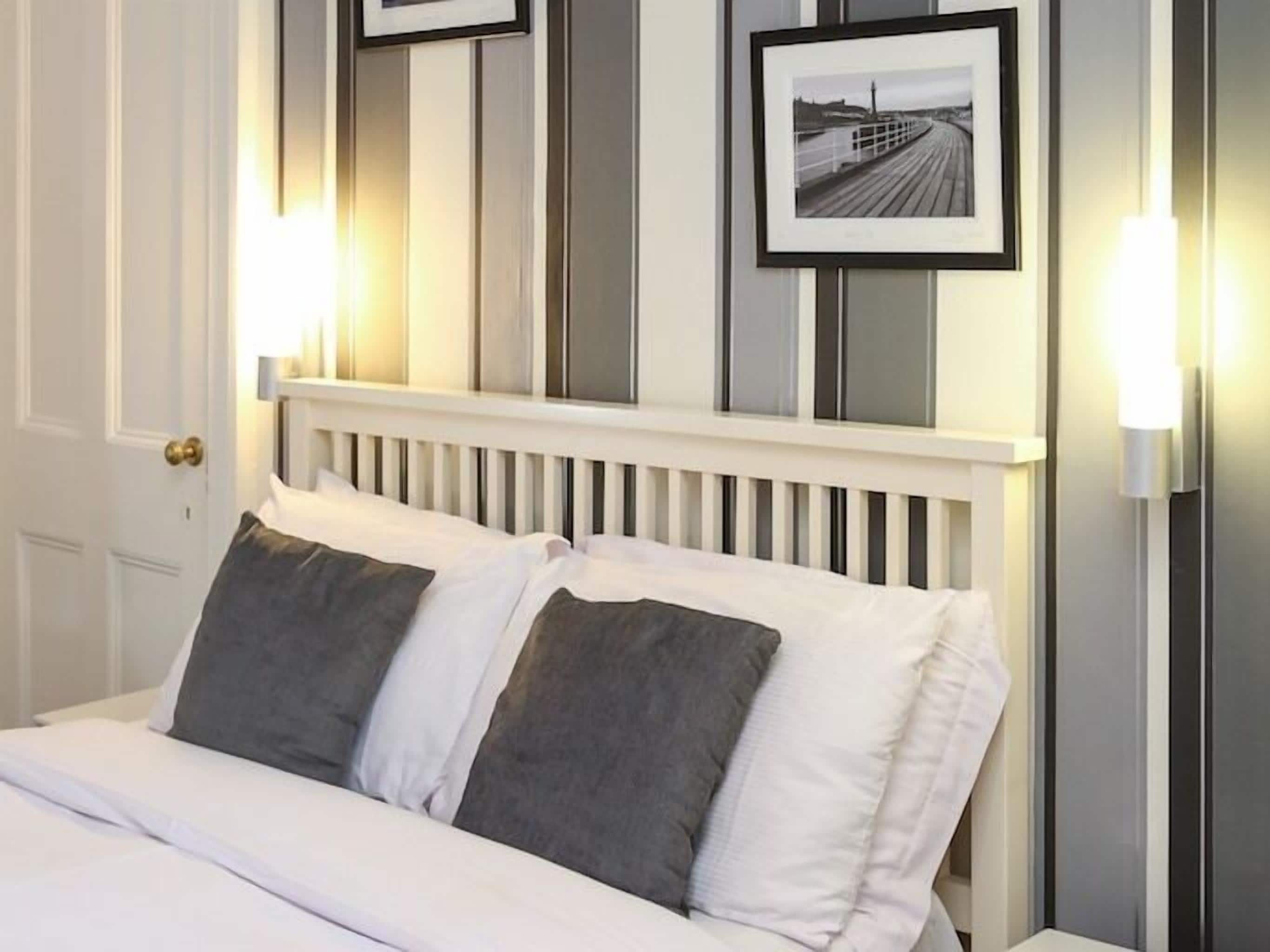 Una cama con ropa blanca y dos almohadas, cabecera blanca y dos cuadros en la pared.