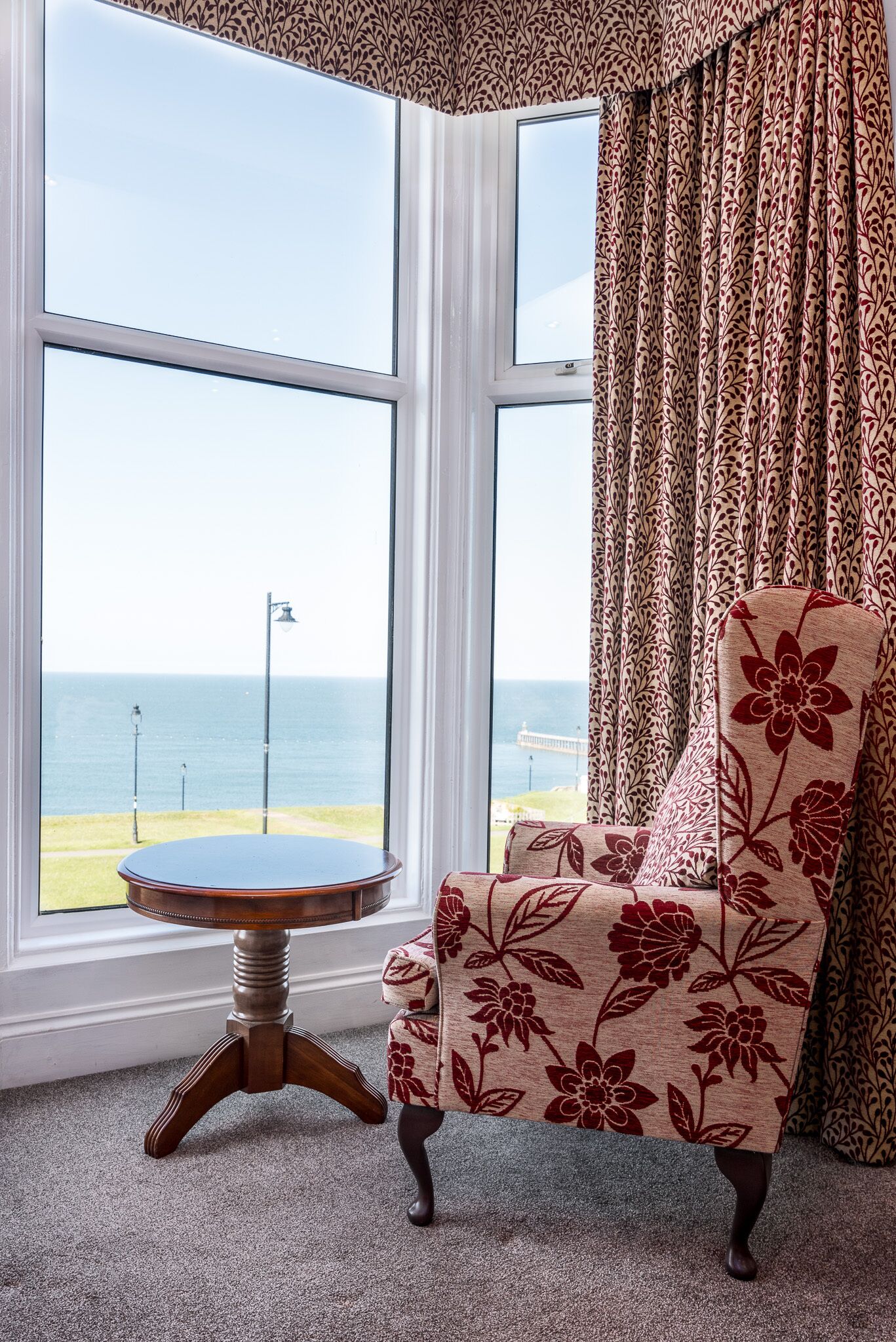 Ein Eckzimmer mit großem Fenster, einem armchair mit Blumenmuster und einem kleinen runden Tisch.