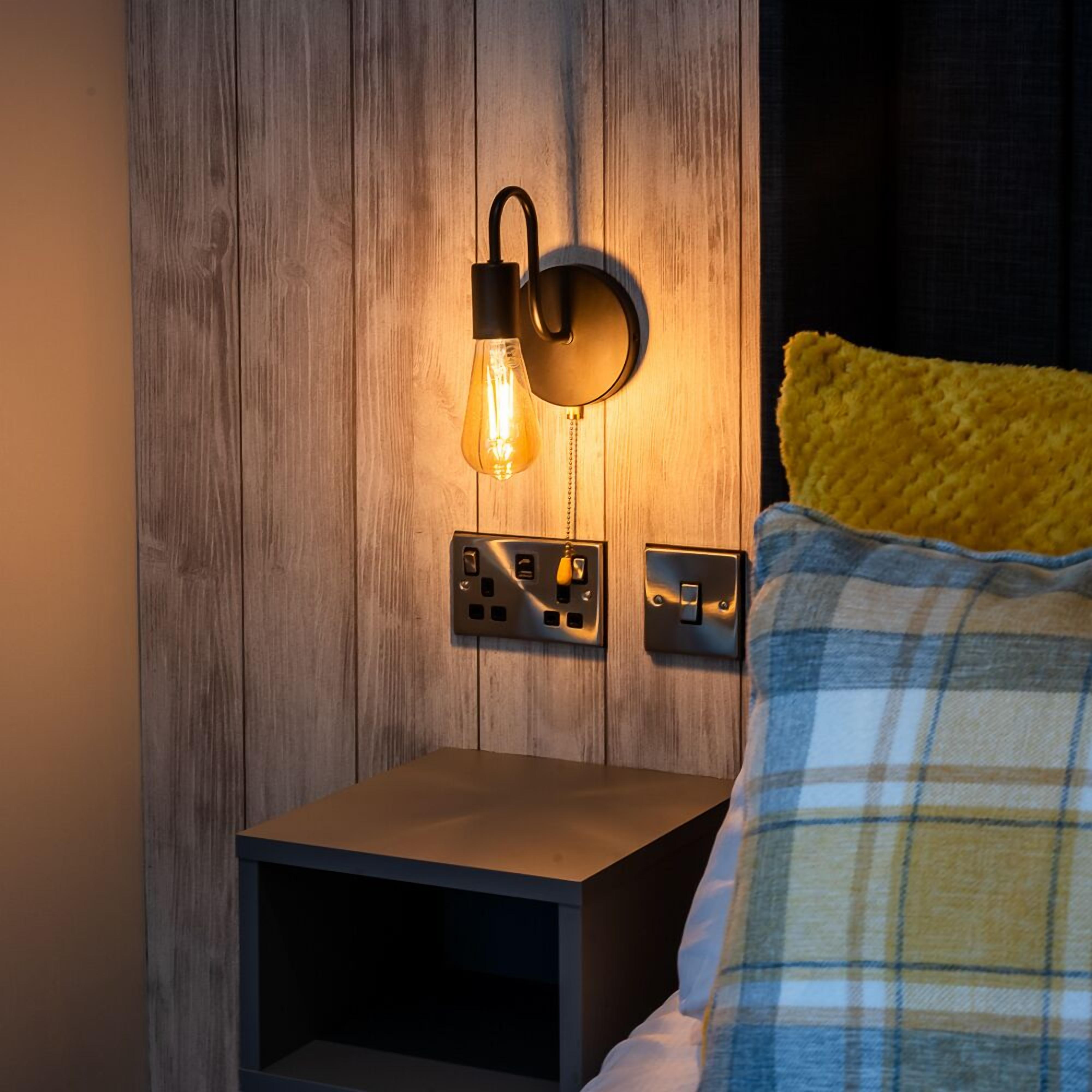 A bed with a yellow pillow and a blue and white checkered blanket, a nightstand with a wall-mounted light fixture, and a wooden wall.