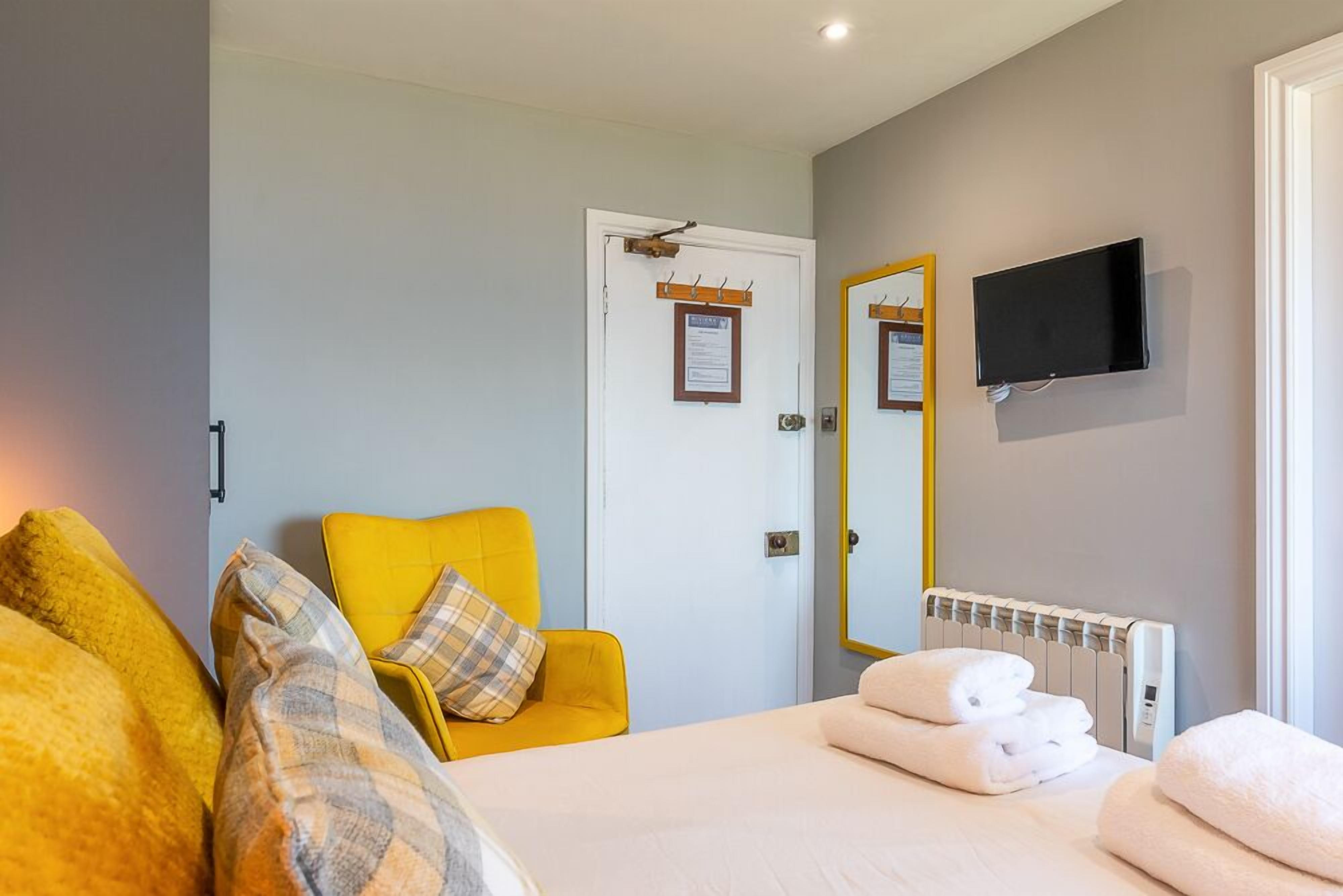 A hotel room with a bed, a yellow armchair, a white bedside table with towels, a mounted TV, and a door with a sign.