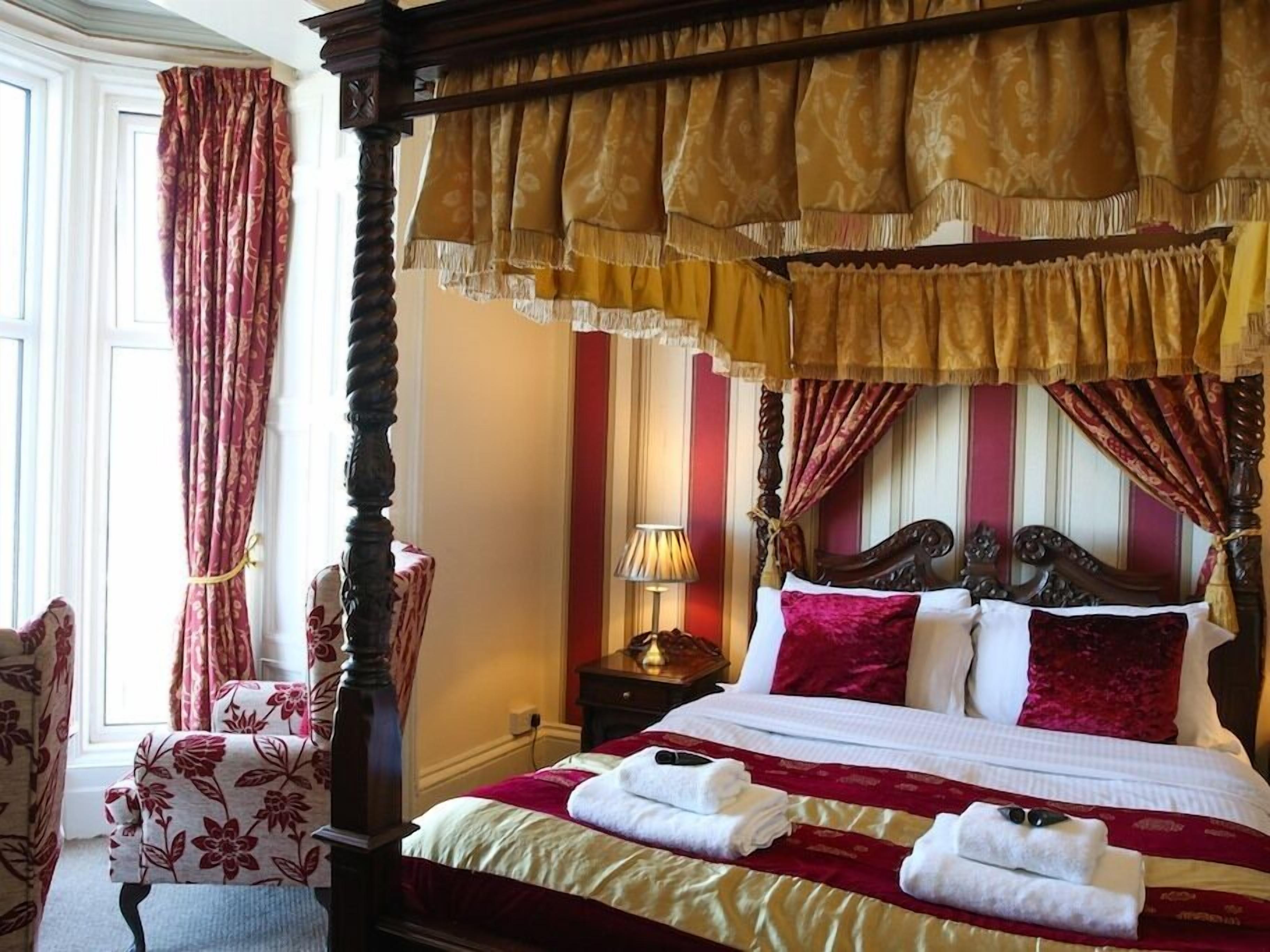 A four-poster bed with a canopy, a chair with floral upholstery, and a bedside table with a lamp.
