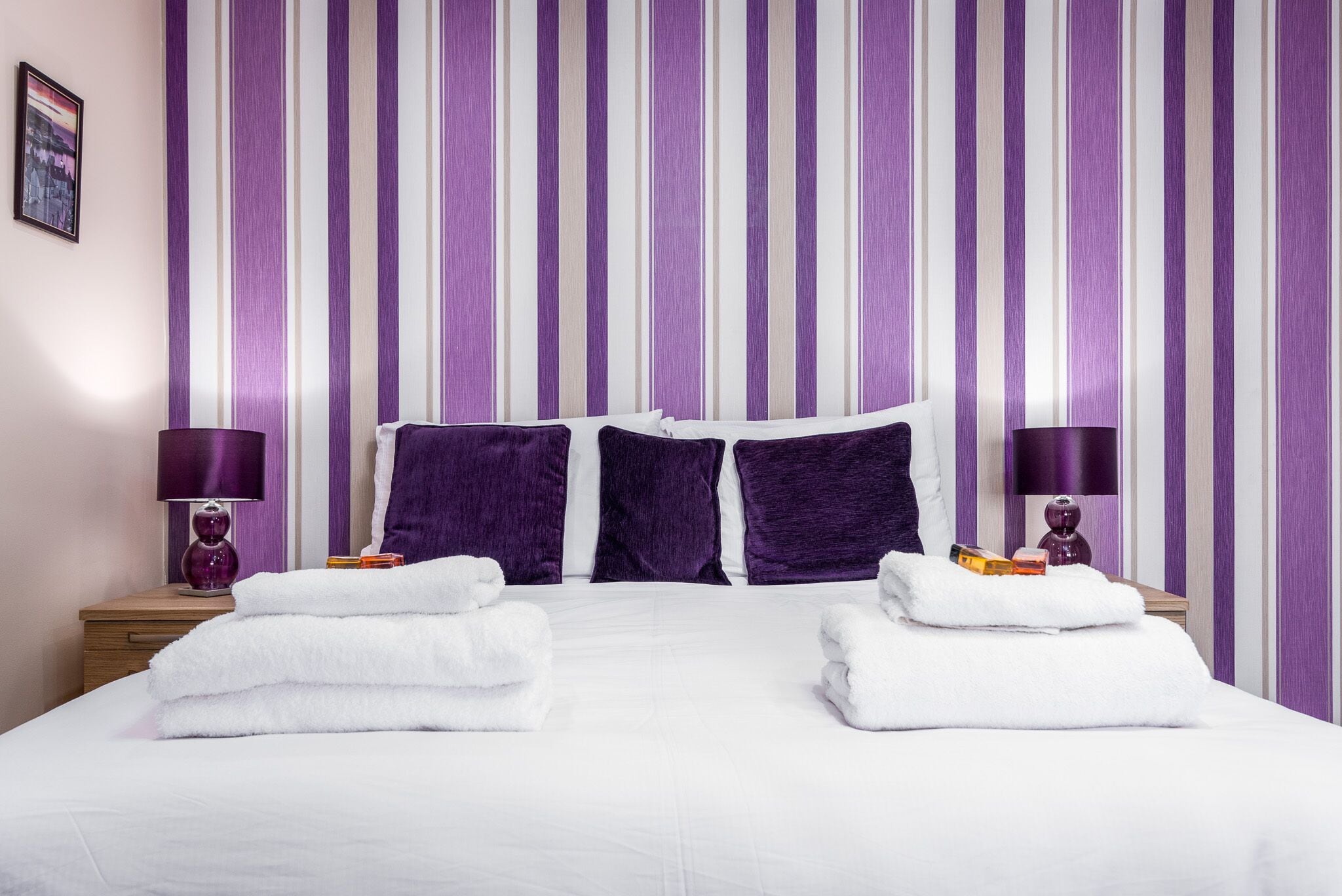 Ett hotellrum med en randig, lila och vit vägg, en snyggt bäddad säng med vita sängkläder, två lila kuddar och två sängbord med lila lampor och en liten prydnadsföremål på varje.