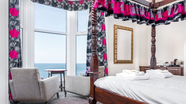 A four-poster bed with a view of the ocean, a chair, a small table, and a mirror on the wall. - The Riviera Guest House (Whitby)