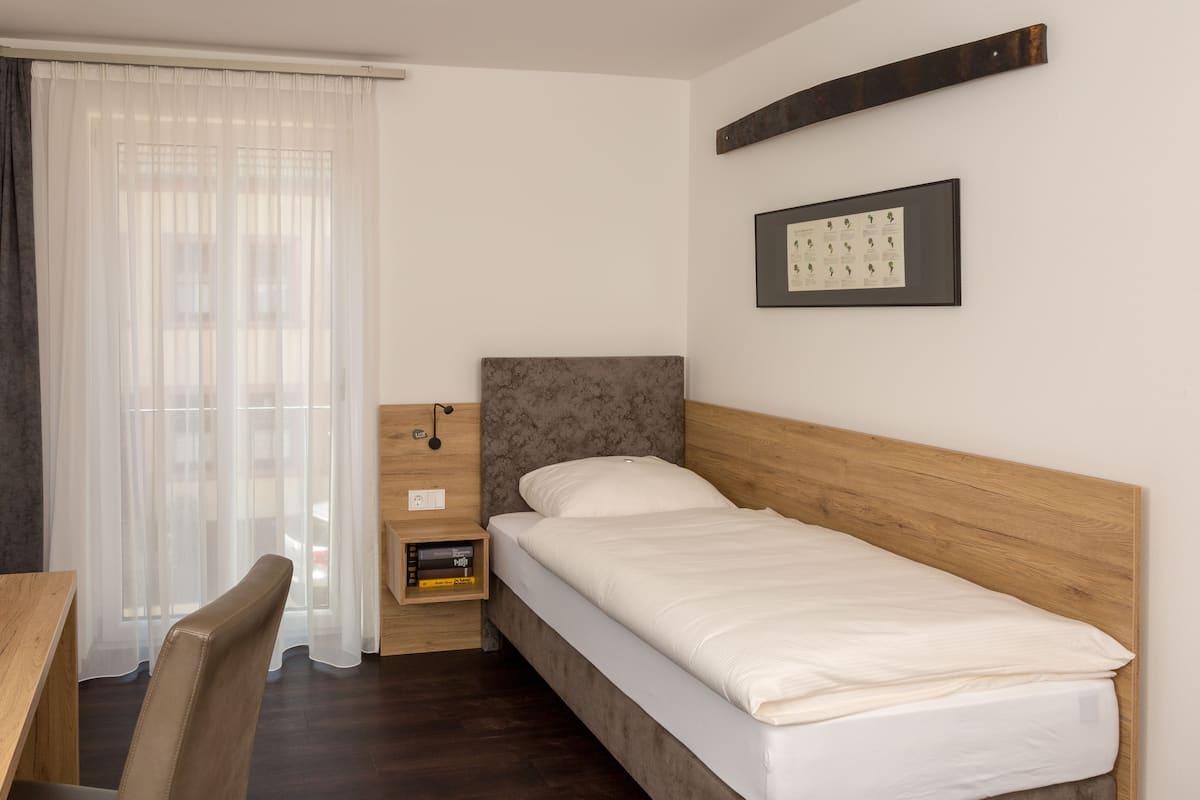 Un dormitorio ordenado con una cama individual, cabecera de madera, mesita de noche con lámpara y ventana con cortinas translúcidas.