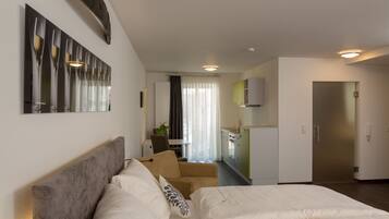 A modern hotel room with a large bed, a sofa, a dining area, and a kitchenette.