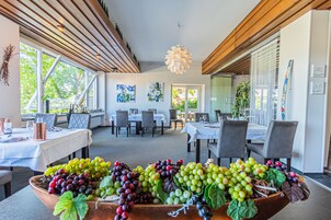 A dining area with tables set for a meal, a large bowl of assorted grapes in the foreground, and a view of the outdoors through large windows.