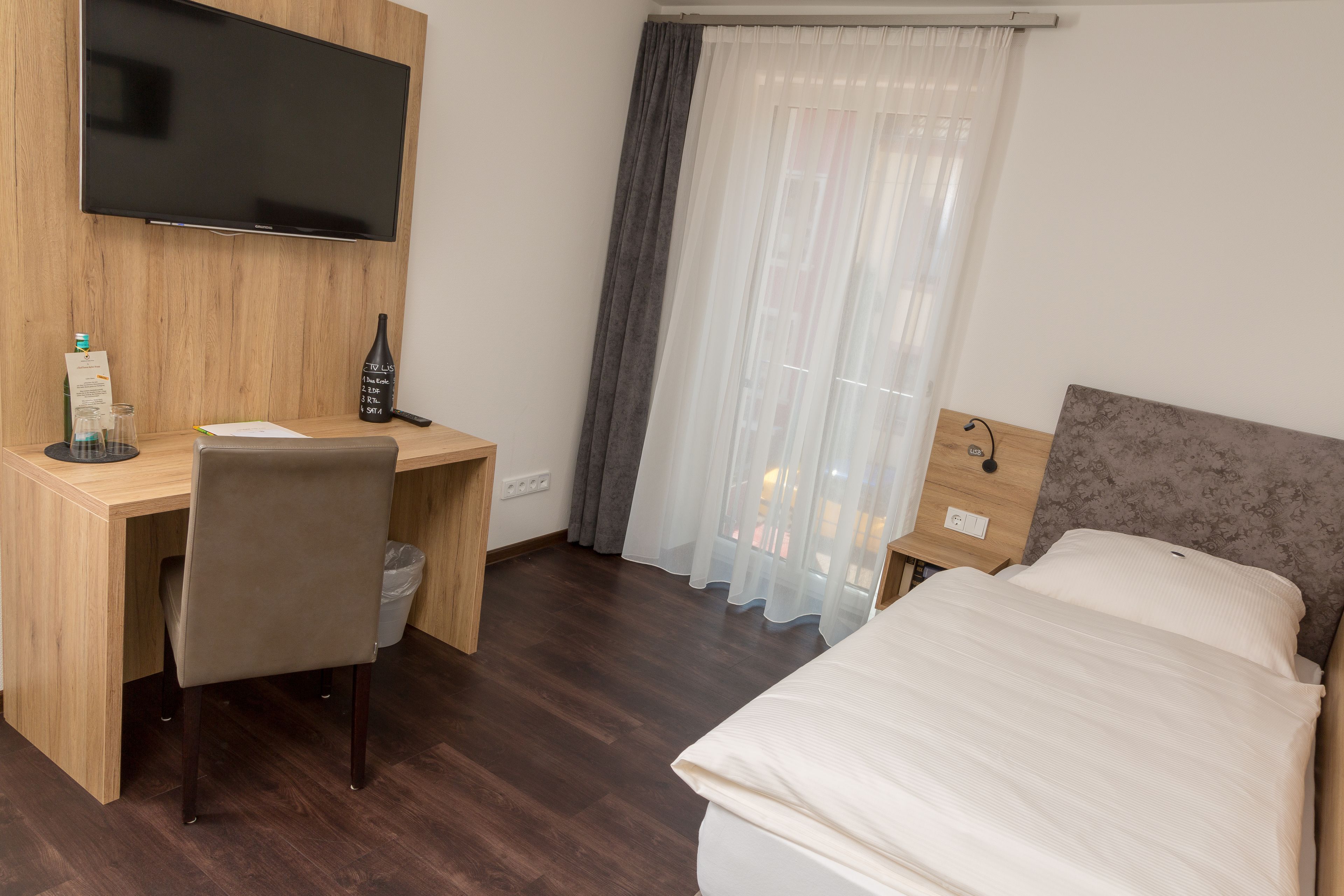 Ein Hotelzimmer mit Bett, Schreibtisch mit Stuhl, Wandfernseher und einem Fenster mit Vorhängen.