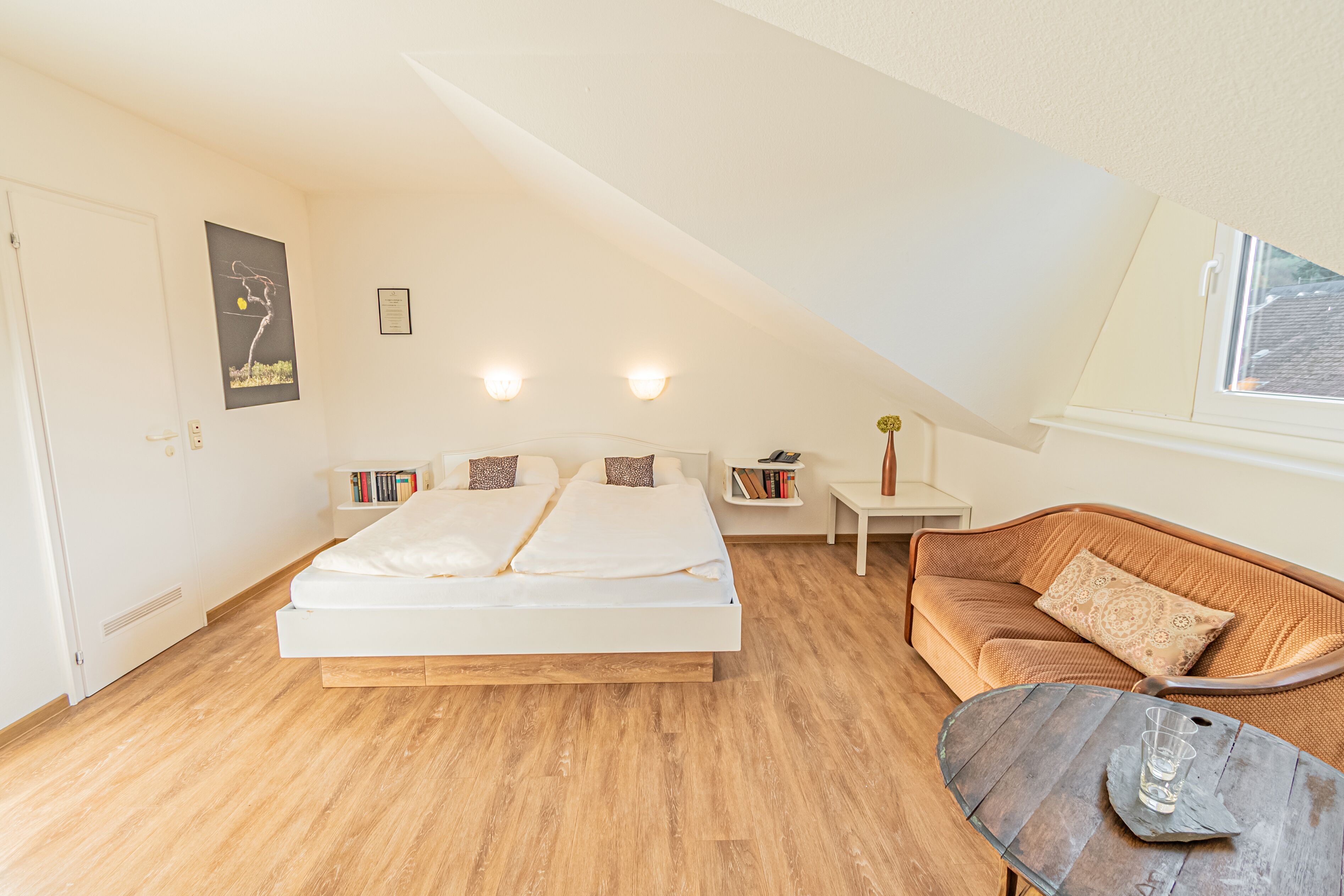 Een slaapkamer met een bed, houten vloer, witte muur, een kleine tafel, een bank en een raam.