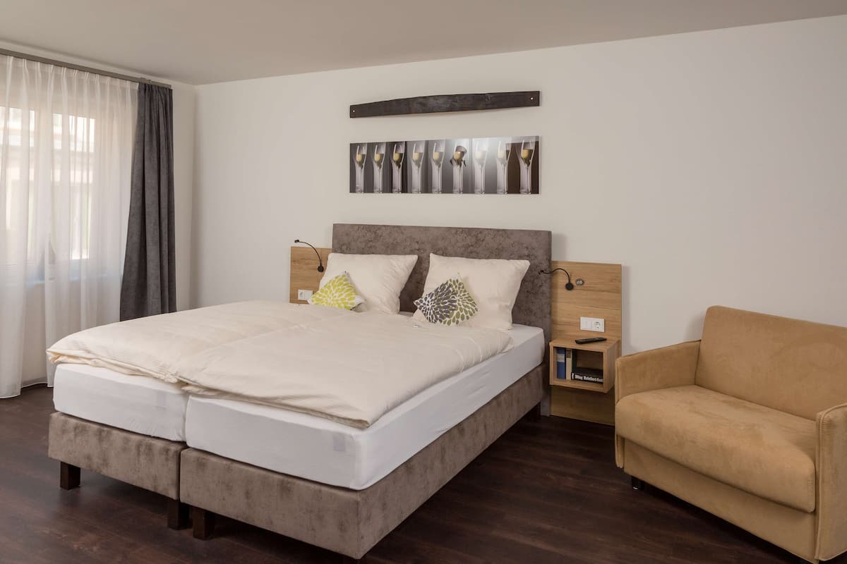 Una cama doble con cabecera, dos mesitas de noche, un sofá marrón, una obra de arte colgada en la pared y una ventana con cortinas.