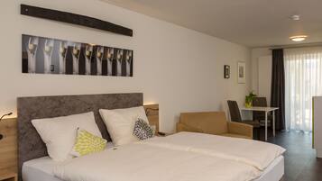 Quarto de hotel com uma cama grande, uma escrivaninha e uma cadeira. Há um quadro na parede e uma janela com cortinas.