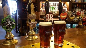 Dos vasos de cerveza sobre la barra de un bar, con grifos identificados como 'Arkells' y 'Wiltshire Gold'.