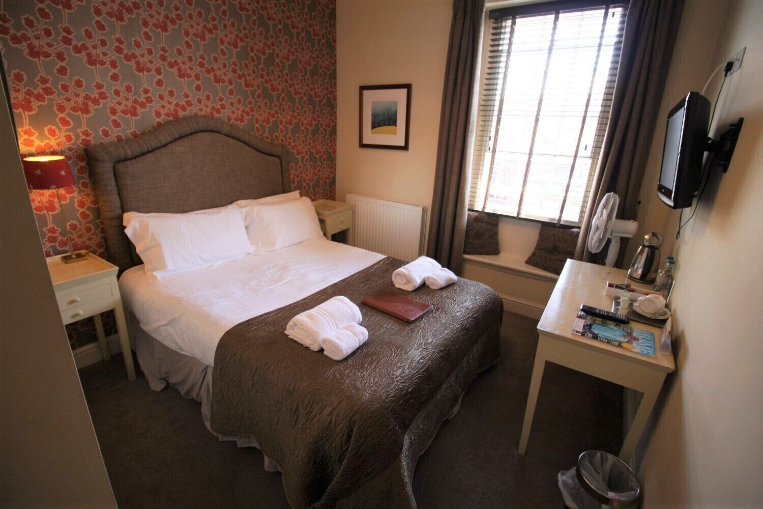 A hotel room with a bed, bedside table, lamp, window, and a desk with a mirror and various items.