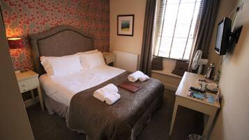 A hotel room with a bed, bedside table, lamp, window, and a desk with a mirror and various items.