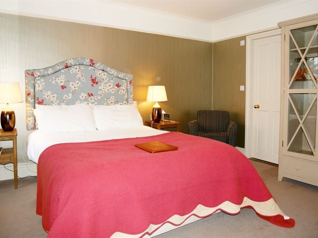 A neatly made bed with a floral headboard, a red bedspread, and a bedside table with a lamp. There is a striped chair and a cabinet with glass doors in the room.