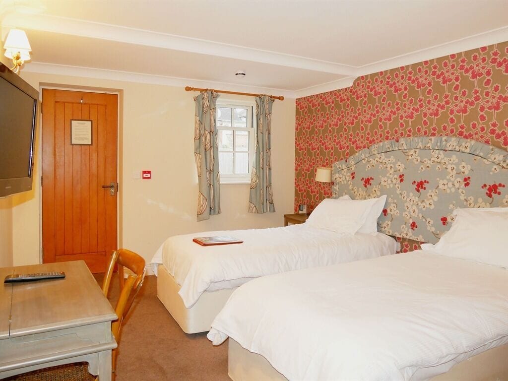 A hotel room with two beds, a wooden door, a desk with a remote control, and a window with curtains.