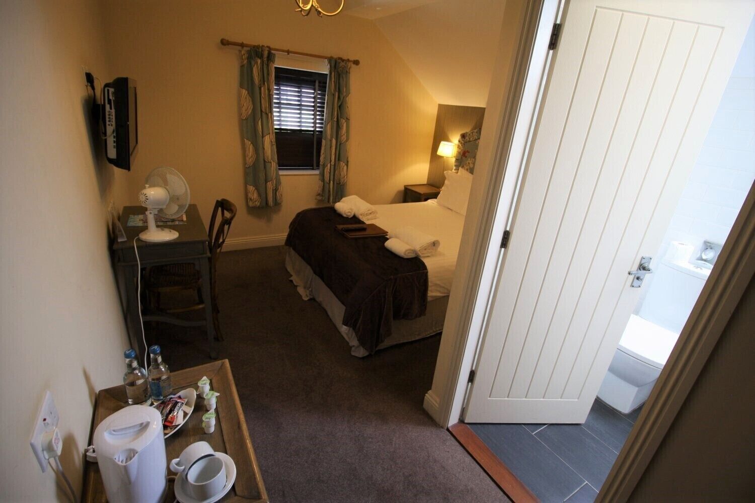 A compact hotel room with a bed, bedside table, fan, and a small desk with a chair. A bathroom is visible through an open door.