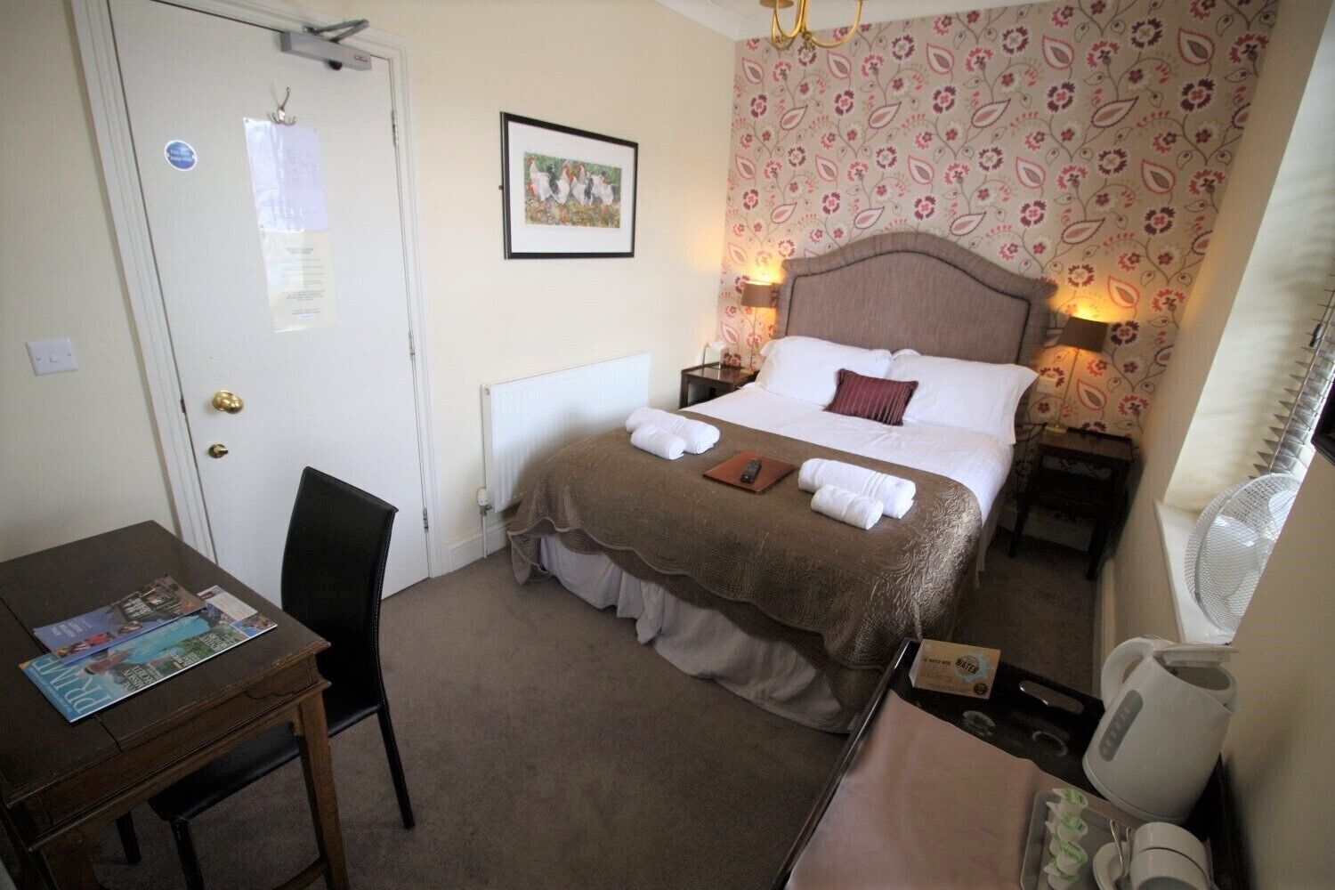 Habitación de hotel con cama, mesitas de noche, escritorio y una silla. Hay un radiador debajo de la cama y una ventana con ventilador.