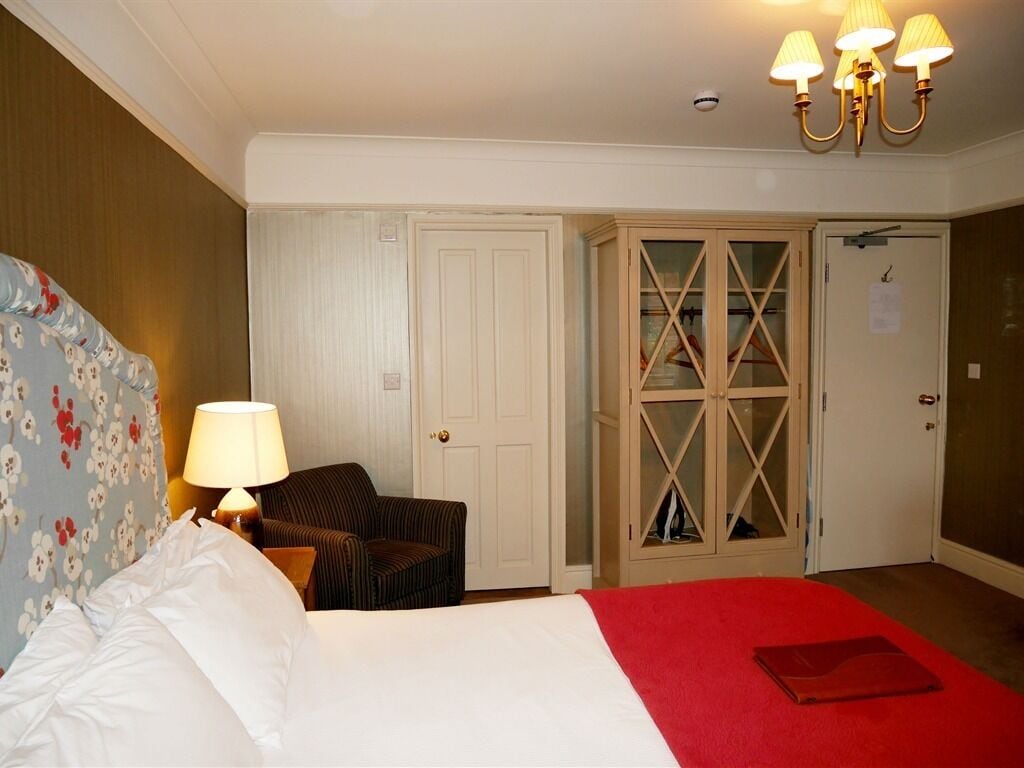 Habitación de hotel con cama, mesita de noche con lámpara, una silla, armario y una puerta con panel de vidrio.