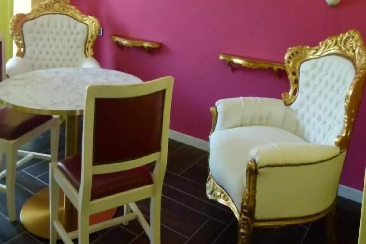 Un comedor con una mesa redonda y dos sillas, ambas con detalles dorados. La pared está pintada de rosa y hay dos apliques.