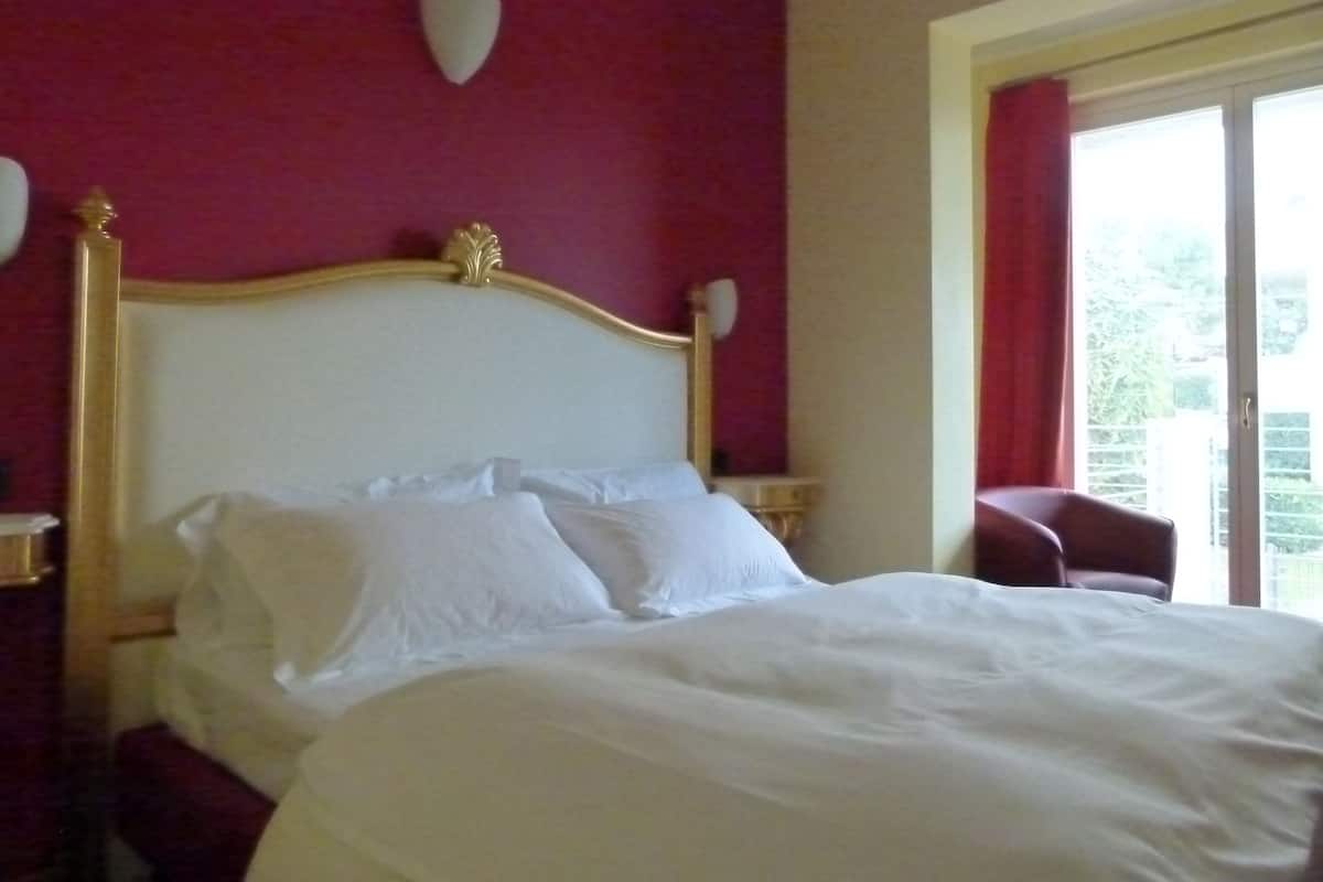 Una habitación de hotel con una cama grande, un cabecero con un escudo decorativo, dos mesitas de noche y una ventana con cortinas.
