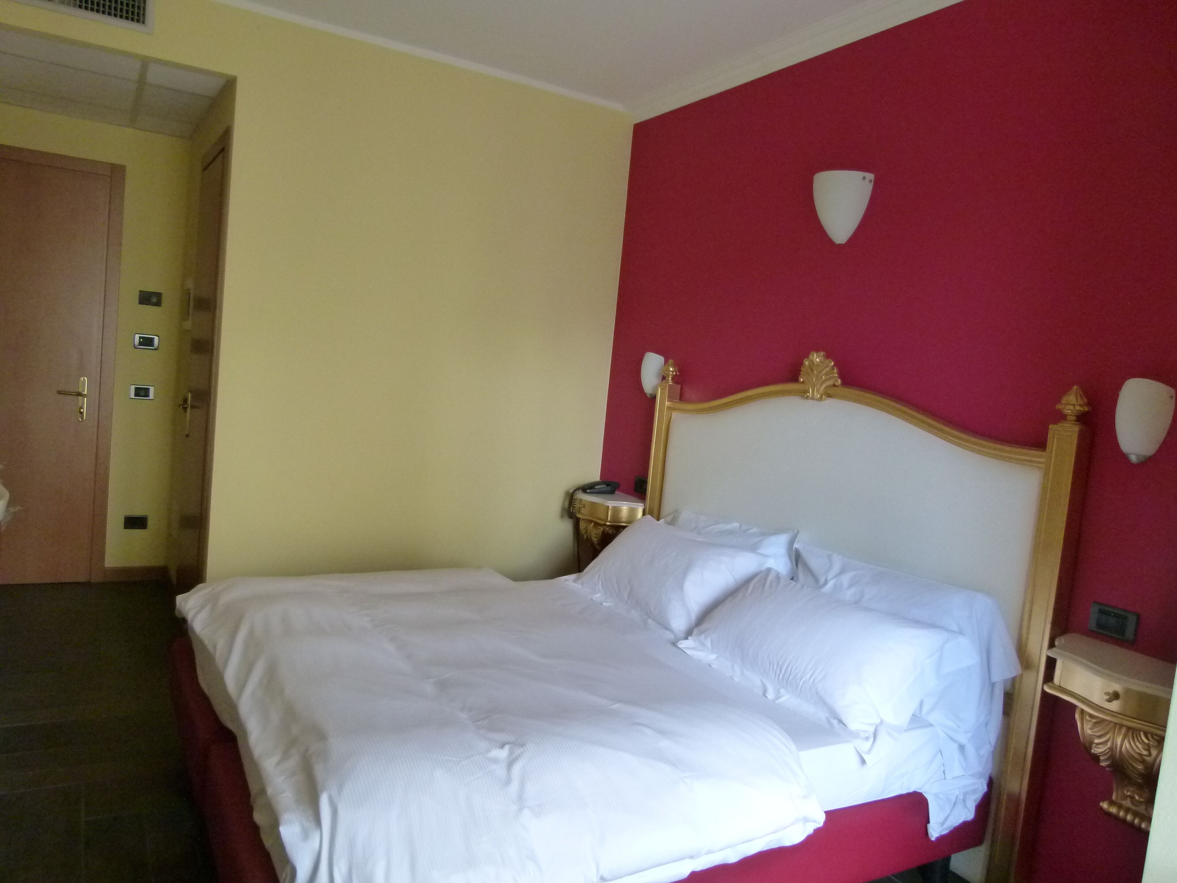 A hotel room with a bed, a nightstand, a wall clock, and a door.