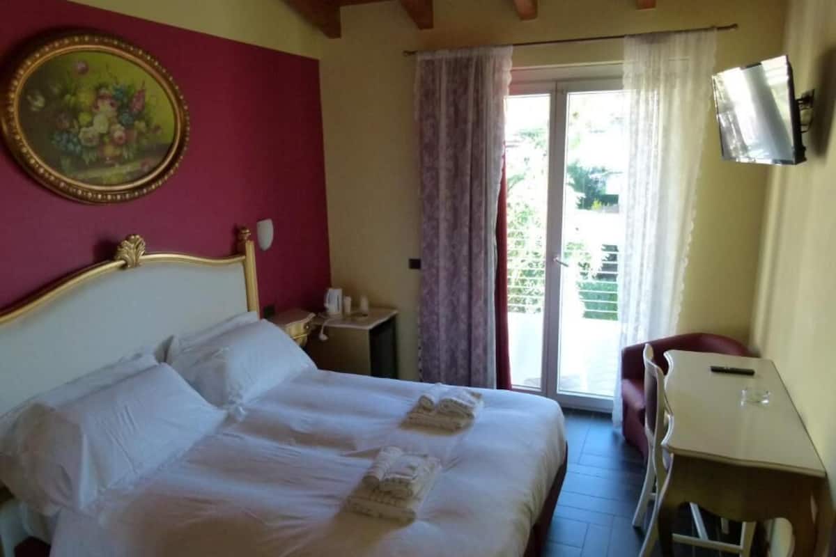 Habitación de hotel con cama, escritorio, silla, televisor y balcón con cortinas.