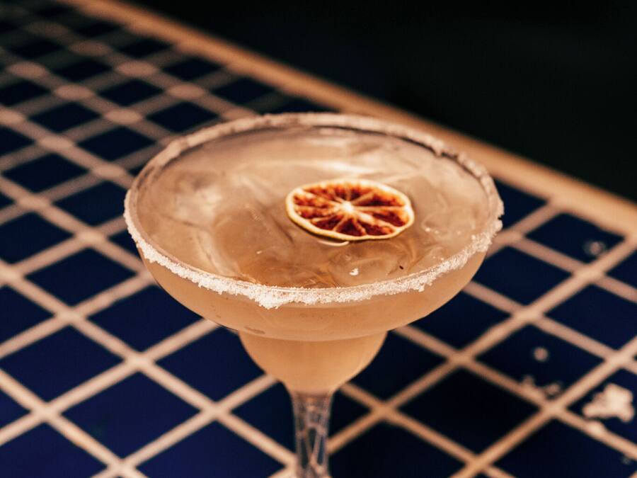 Un verre à cocktail avec un bord saupoudré de sel et une tranche d’orange séchée, posé sur une surface recouverte de carreaux bleus.