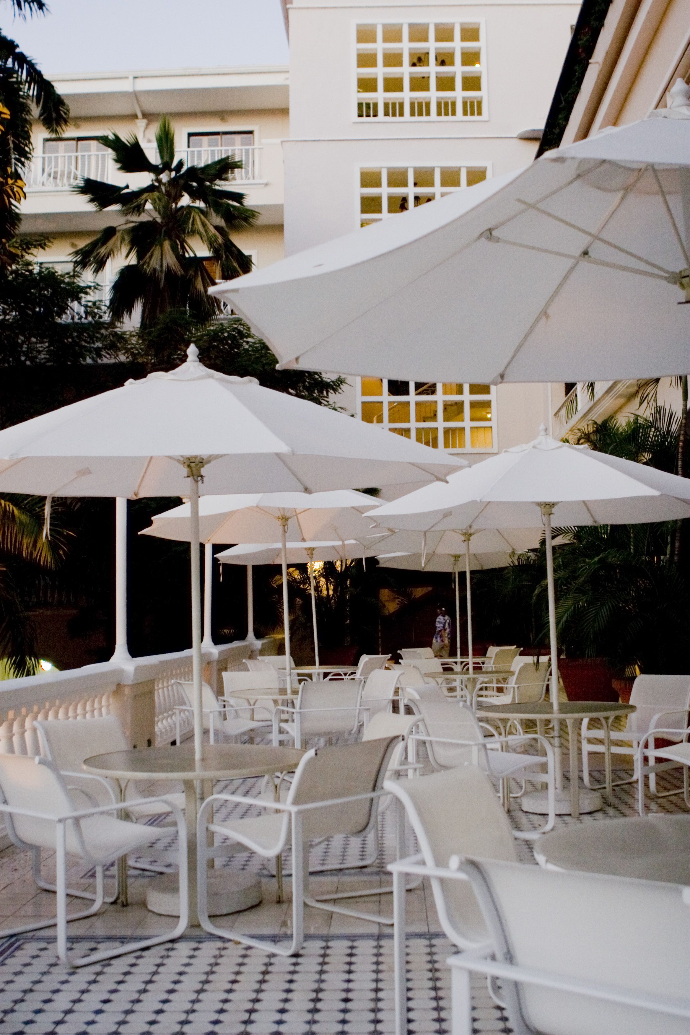 Una terraza con mobiliario exterior blanco y sombrillas, rodeada por un edificio con balcones y palmeras.