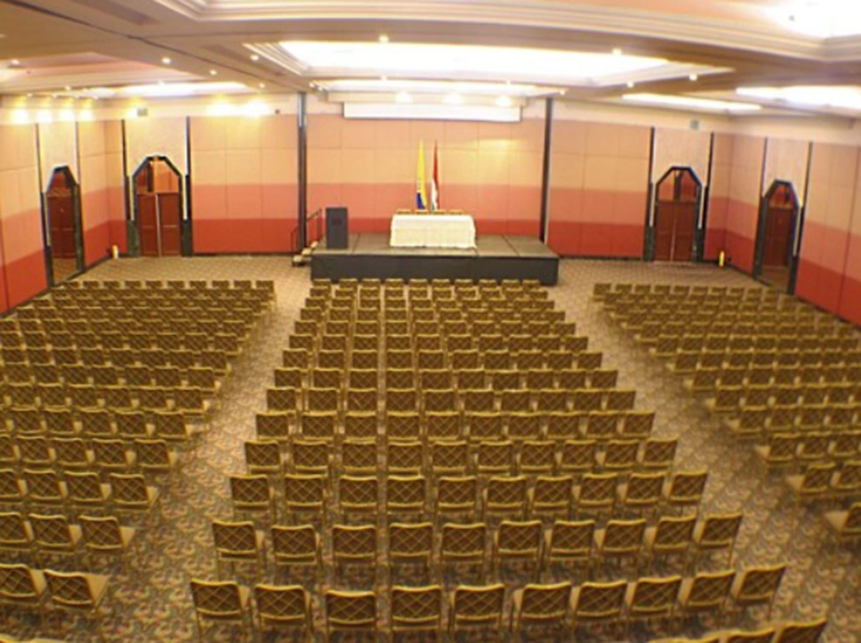 Une salle de conférence avec des rangées de chaises, une scène avec un pupitre et des drapeaux.