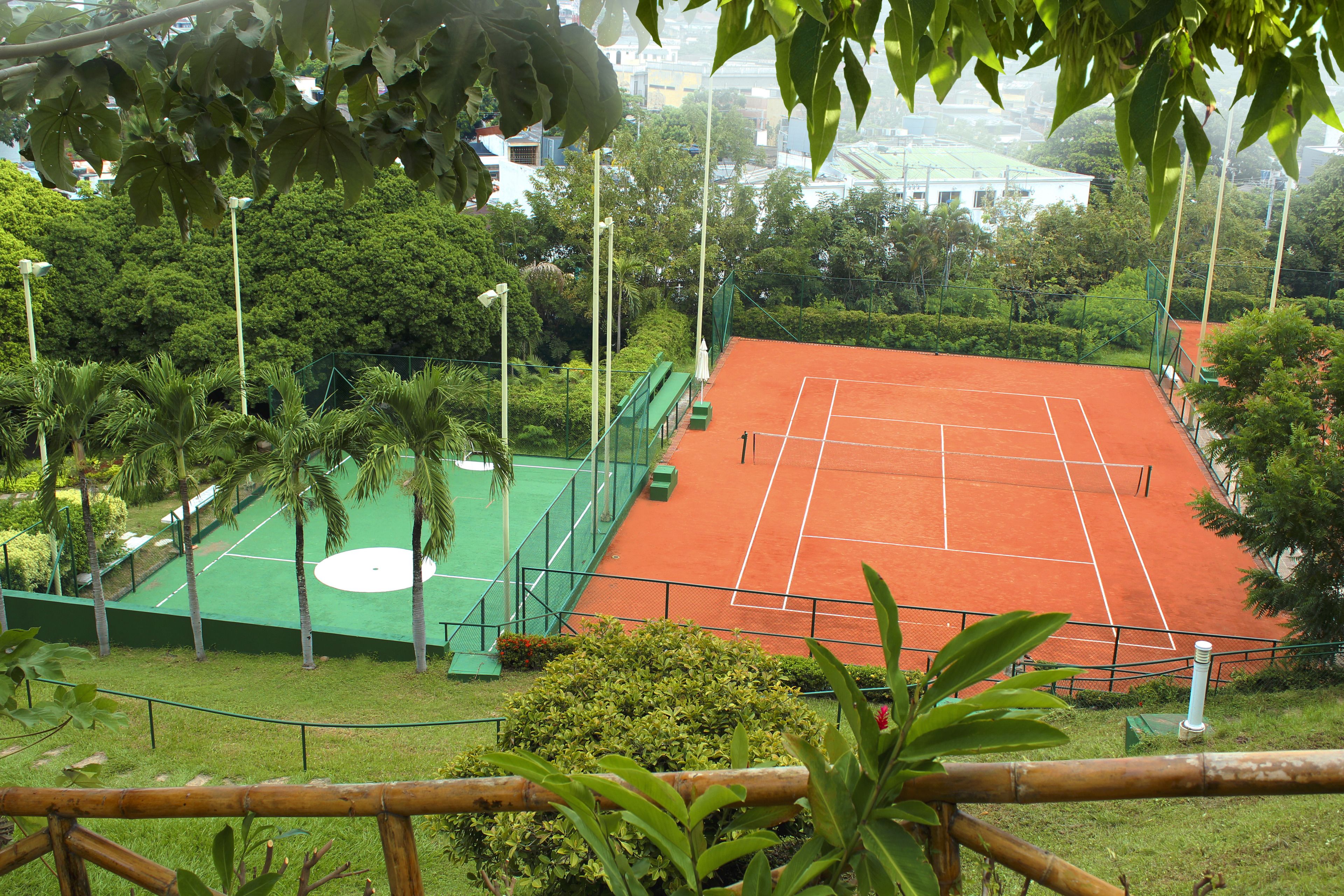 A tennis court surrounded by greenery and a building in the background.
