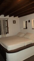 Un dormitorio con estructura de cama de madera, ropa de cama blanca, una mesita de noche pequeña con una lámpara y una ventana con cortina.